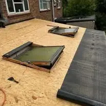 New timber boards on a flat roof with two rooflights in place and a roll of covering at the edge